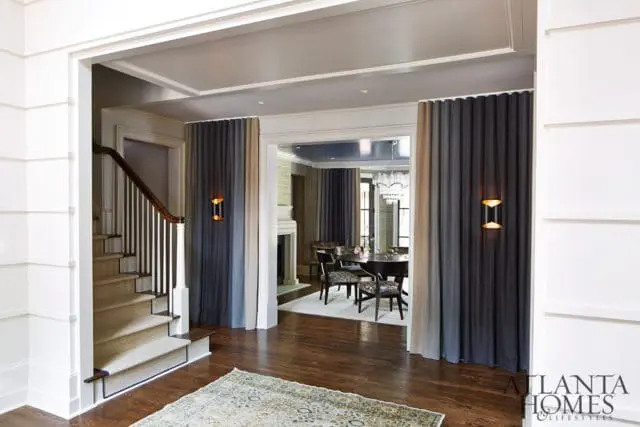 Foyer with staircase, dark wood floors, and ombre curtains leading to a dining room with a chandelier and fireplace.