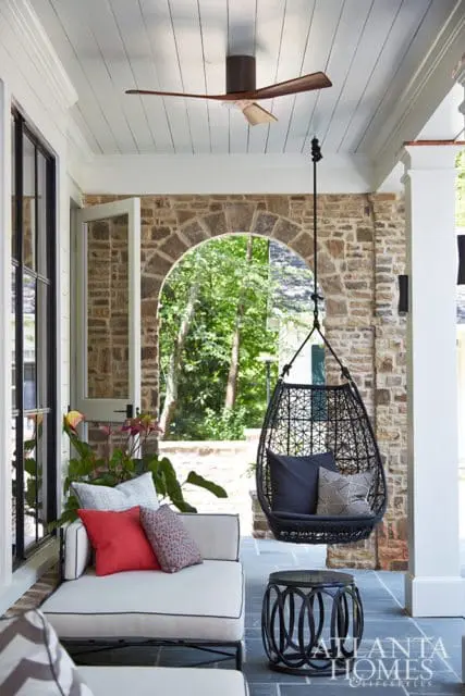 Covered porch with stone archway, white ceiling fan, white outdoor sofa with red and patterned pillows, and a black hanging egg chair with cushions.