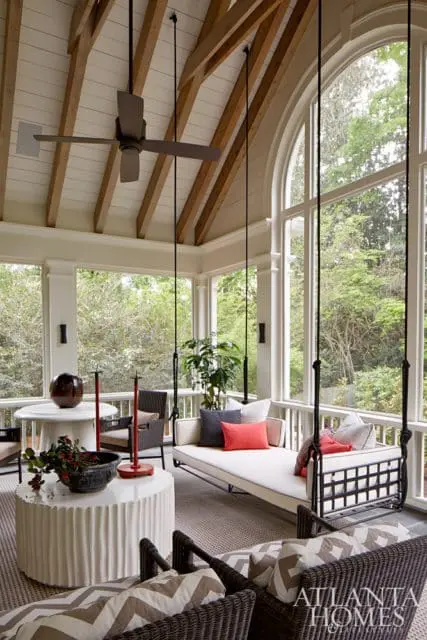 Sunroom with a hanging daybed, wicker chairs, a round table, and a large arched window overlooking trees.