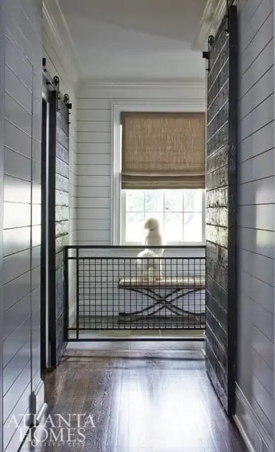 Hallway with white shiplap walls, dark wood barn doors, and a black metal railing. A white dog sits on a bench by a window with a linen Roman shade.