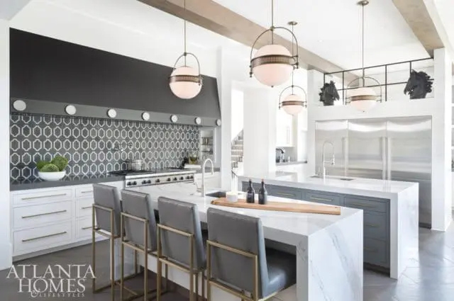 Modern white kitchen with geometric tile backsplash, stainless steel range, marble island with gray cabinetry, and globe pendant lights.
