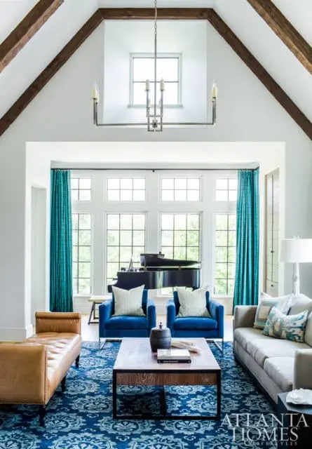 Living room with vaulted ceiling, exposed wooden beams, and a large window wall featuring a grand piano. Furnishings include blue armchairs, a tan leather bench, a grey sofa, and a patterned blue rug.