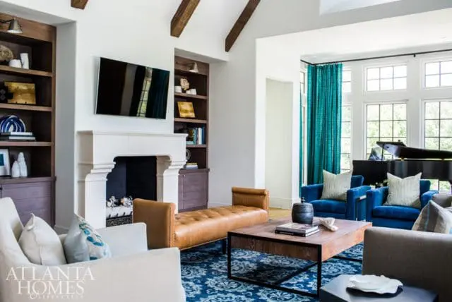 Living room with white fireplace, built-in dark wood shelves, tufted leather bench, blue patterned rug, and blue armchairs facing a grand piano.
