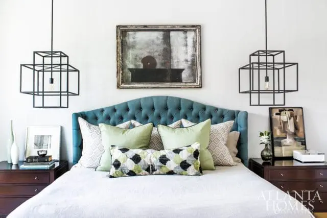 Bedroom with a teal tufted headboard, white linen bedding, sage green and patterned throw pillows, and geometric black pendant lights.