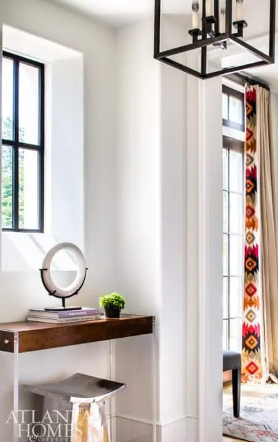 Entryway with a narrow wood console table topped with a circular sculpture, books, and a small plant. A metal stool sits below. Geometric patterned curtains hang near a window.