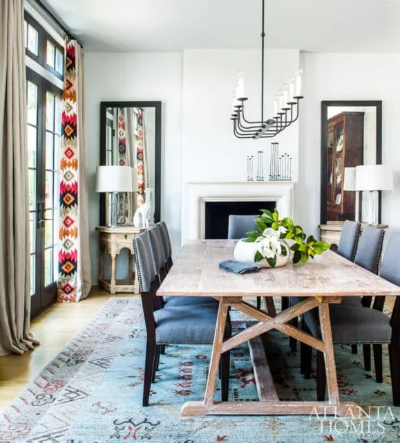 Dining room with light wood table, upholstered gray chairs, fireplace, and patterned curtains.