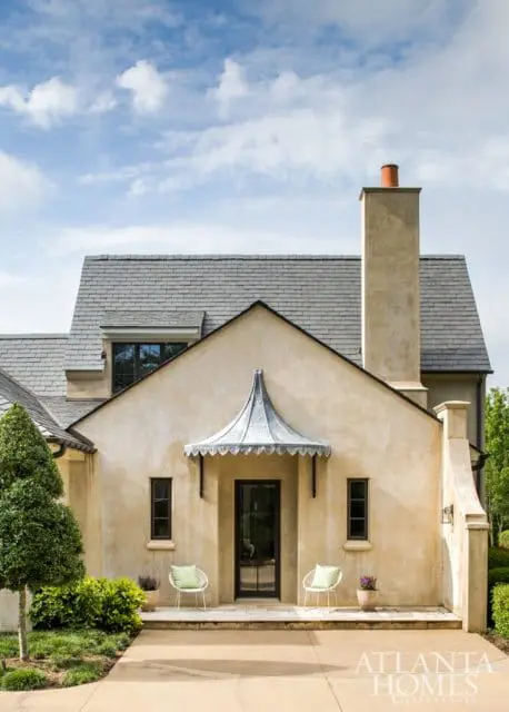 Stucco house with a gray slate roof, a tall chimney, and a decorative metal awning over the front door. Two white chairs with green cushions flank the entrance.