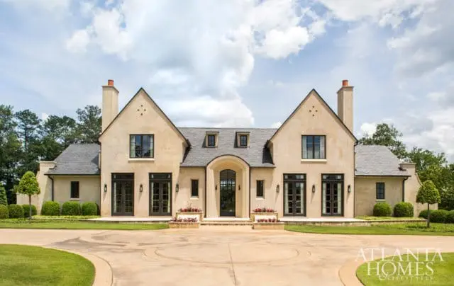 Large stucco house with dark-framed windows, multiple gabled roofs, and symmetrical design. A circular driveway leads to the arched entryway with double doors.