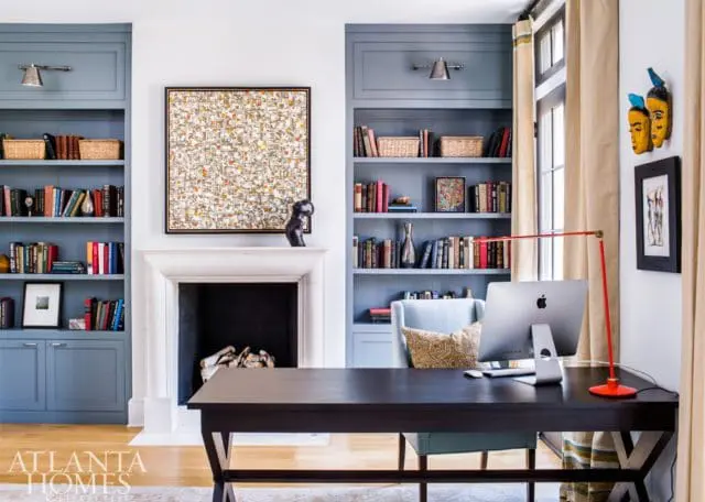 Home office with blue built-in bookshelves flanking a stone fireplace, a dark wood desk with an Apple computer, and a red desk lamp.
