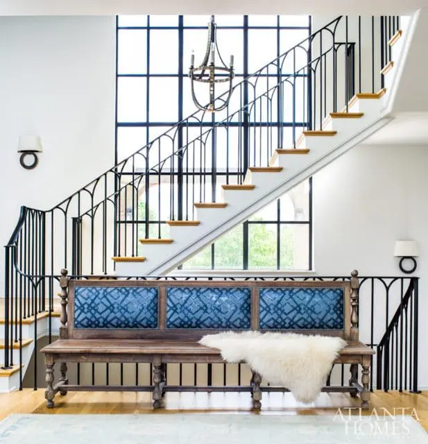 Entryway with a wooden bench upholstered in blue patterned fabric, a white sheepskin throw, and a grand staircase with black metal railings and a large grid window.