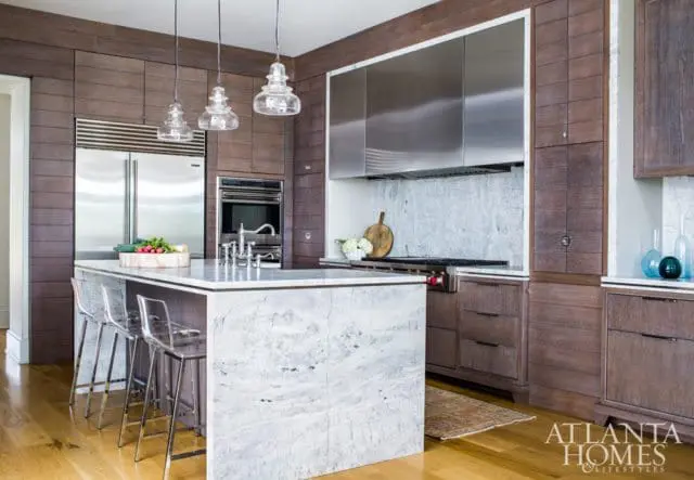 Modern kitchen with dark wood paneled walls, a large marble island with clear stools, stainless steel appliances, and glass pendant lights.