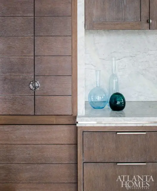 Dark wood cabinetry with horizontal plank detailing and a decorative silver pull, next to a marble backsplash and countertop holding three glass vases.
