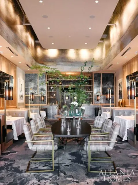 Restaurant dining room with plush booth seating, oval tables set with white linens, and plaid upholstered chairs with gold frames. A large centerpiece of greenery and glass vases sits on a round table.