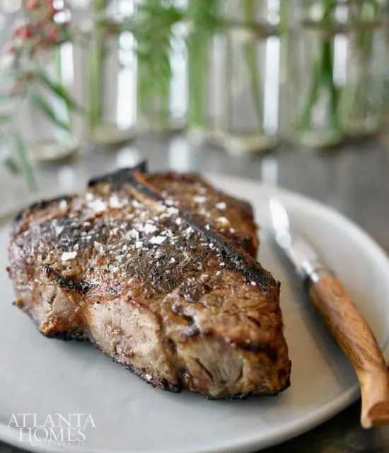 Grilled T-bone steak seasoned with coarse salt, served on a light gray plate with a steak knife