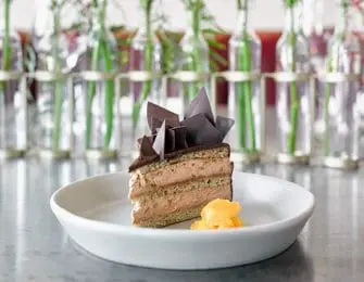 Slice of layered chocolate cake with chocolate shards and a dollop of orange compote on a white plate, with a blurred background of glass vases holding greenery and red liquid.