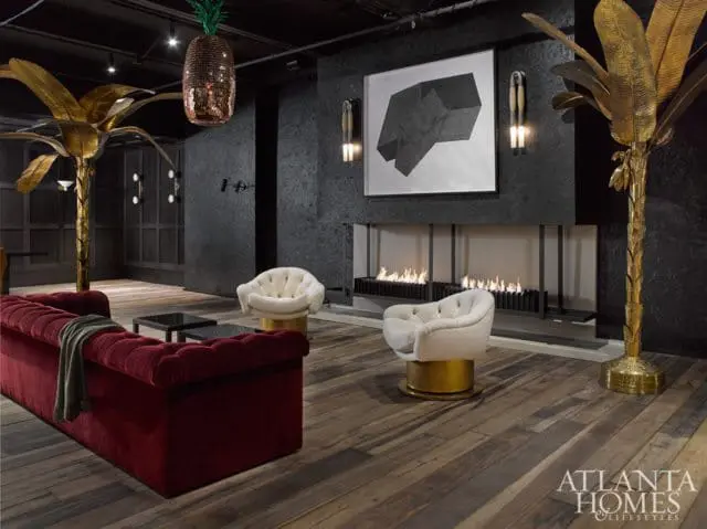 Dark gray living room with a tufted burgundy sofa, two cream swivel chairs with gold bases, a modern fireplace with linear flames, and brass palm tree floor lamps.