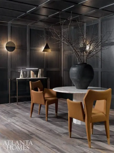 Dark wood paneled dining room with a marble top table, two tan upholstered chairs, and a large black vase with bare branches.