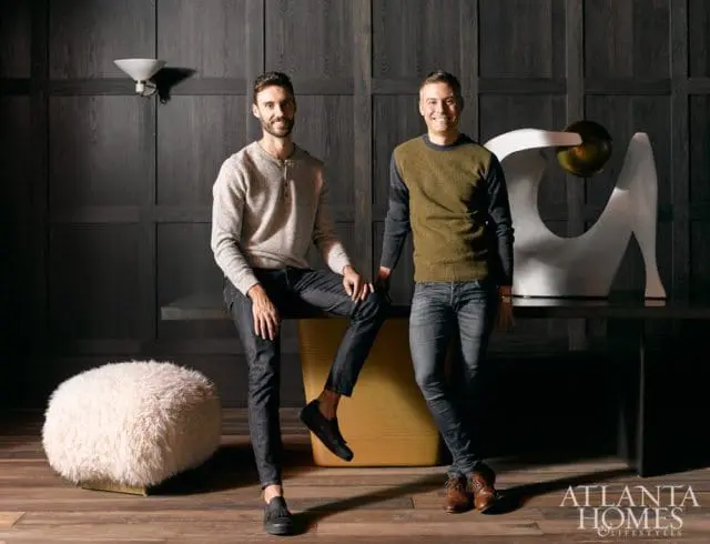 Two men posing in a room with dark wood paneled walls. One man sits on a bench, the other stands. A white sculpture and a fluffy ottoman are visible.