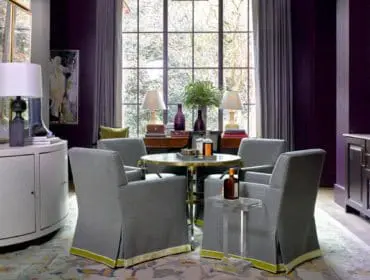 Dining area with four gray upholstered chairs around a round metal table, dark purple walls, and a large window overlooking trees.