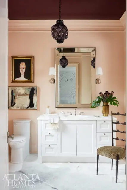 Peach-toned bathroom with a white vanity, marble floor, and two framed artworks above a toilet. A dark purple lantern hangs from the ceiling.