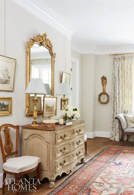 Entryway with a distressed antique dresser topped with lamps and flowers, a gilded mirror, and a patterned runner rug.