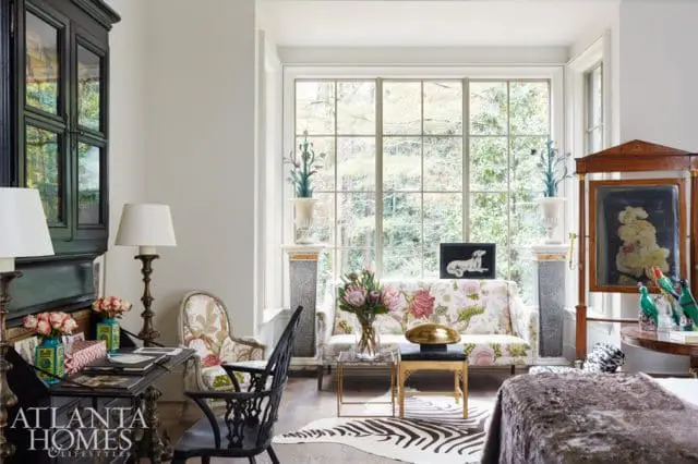 Living room with floral sofa, dark wood desk, ornate chair, and large window overlooking greenery. Atlanta Homes & Lifestyles logo in bottom left.
