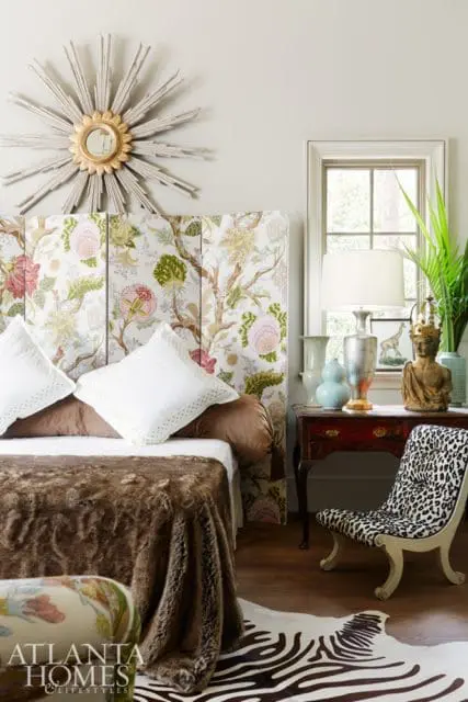 Bedroom with a floral upholstered headboard, white pillows, a brown fur throw, a sunburst mirror, and a leopard print chair next to a dark wood table with decorative objects.