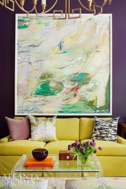 Living room with a bright yellow sofa, abstract art, and a clear acrylic coffee table with books and flowers.