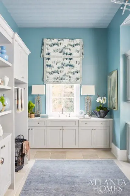 Blue laundry room with white cabinetry, a patterned roman shade over the window, and a blue textured rug on stone tile flooring.