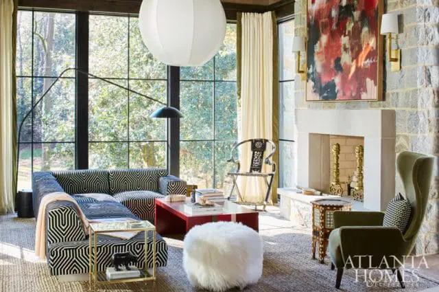 Living room with a black and white geometric patterned sectional sofa, red coffee table, white fur ottoman, and a green wingback chair. A stone fireplace with abstract art above is visible.