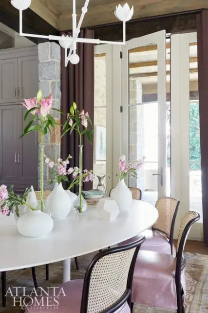 Dining room with a white oval table, cane-backed chairs with pink upholstery, and a modern white chandelier. Pink lilies in glass vases adorn the table.