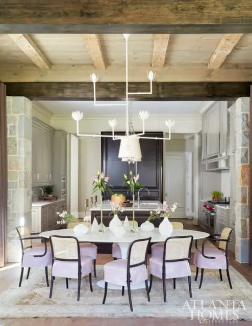 Dining room with oval table, cane-backed chairs with pink upholstery, and a white chandelier. Kitchen visible in background with gray cabinetry and stone accents.