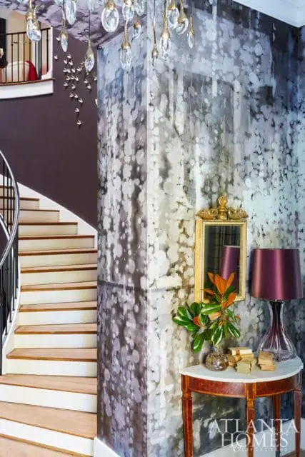Foyer with a curved staircase, dark purple walls, and a column covered in abstract patterned wallpaper. A console table holds a lamp, books, and a plant.