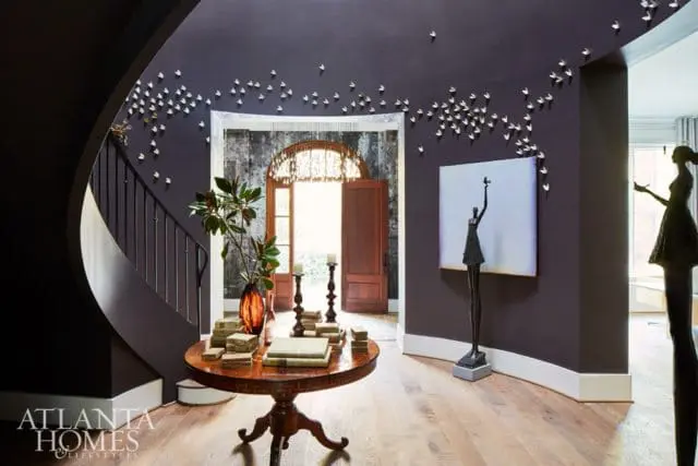 Dark purple entryway with a curved staircase, a round wooden table with books, and wall art of white birds and sculptures.