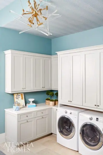 Laundry room with light blue walls, white shaker cabinets, white quartz countertops, two white front-load washers, and a modern gold and glass chandelier.