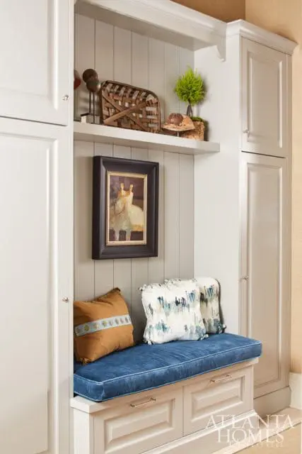 Built-in bench with blue velvet cushion and decorative pillows, flanked by white paneled cabinets and a shelf displaying decorative objects and a painting.