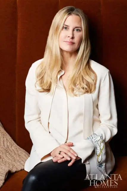 Woman with blonde hair wearing a cream blouse and black pants, seated against a rust-colored upholstered wall.
