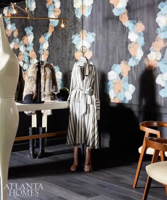 Dark wood floor in a room with a wall featuring abstract cloud-like wallpaper in blue, peach, and white. A console table displays a fur coat, hat, and wine glass, with a striped dress on a mannequin.