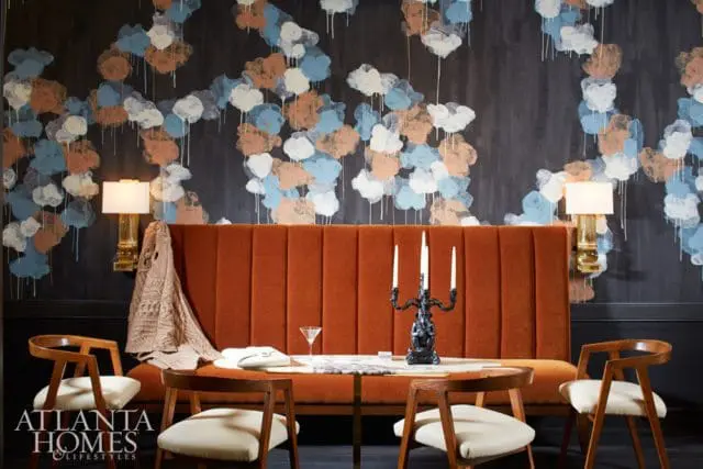 Dining area with a burnt orange tufted banquette, dark wood chairs with cream upholstery, and a dark wall with a painterly cloud-like wallpaper in blue, white, and rust tones.