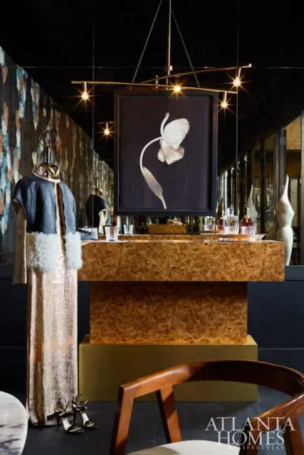 Interior scene with a burl wood console table topped with glassware and a framed botanical print. A dress hangs on a rack to the left, and a wooden chair is in the foreground.