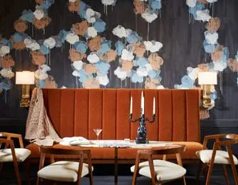 Dining area with a burnt orange tufted banquette, wooden chairs, and a dark wall with abstract blue, white, and brown cloud-like wallpaper.