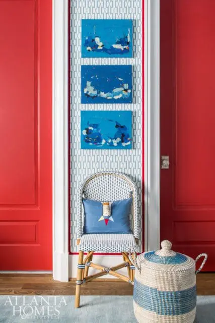 Hallway with bright red doors, blue and white patterned wallpaper, three abstract blue paintings, a white wicker chair with a blue pillow, and a woven basket.