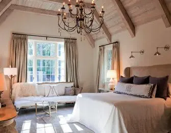 Master bedroom with vaulted wood-beamed ceiling, large window with draped curtains, king-size bed with plush headboard, and ornate black chandelier.