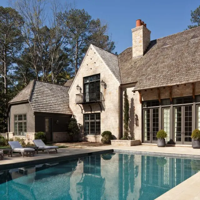Stone mansion with a shingled roof, dark-framed windows, a small balcony, and a large swimming pool in the foreground.