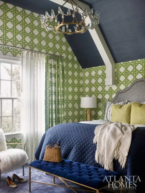 Bedroom with navy blue ceiling, green lattice wallpaper, and a large bed with a navy blue quilted comforter and yellow pillows. A white shag chair and a blue velvet bench are in the foreground.