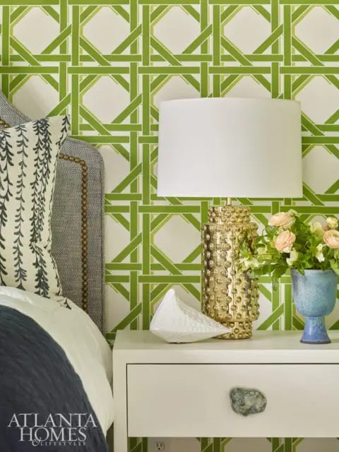 Bedroom with green lattice wallpaper, upholstered headboard, white nightstand with gold lamp, white bird sculpture, and blue vase with flowers. Atlanta Homes & Lifestyles logo in lower left corner.