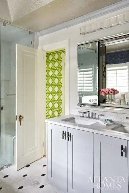 Light gray double vanity with marble countertop and silver hardware, adjacent to a glass-enclosed shower and a doorway with green geometric wallpaper.