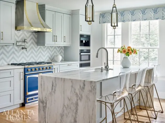 Light gray kitchen with a blue range, marble island, chevron tile backsplash, and brass accents.