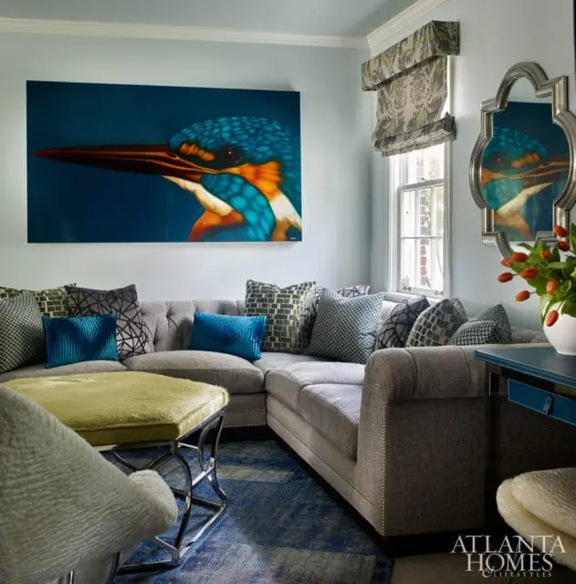 Light blue living room with a large kingfisher painting, gray sectional sofa with patterned pillows, lime green ottoman, and a blue rug.