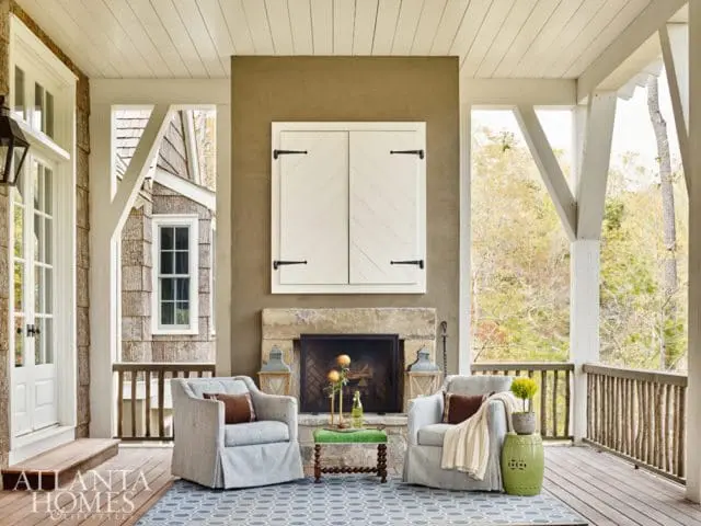 Covered porch with a stone fireplace, two upholstered chairs, and a blue patterned rug. White shutters adorn the fireplace, and a wooden railing lines the porch.